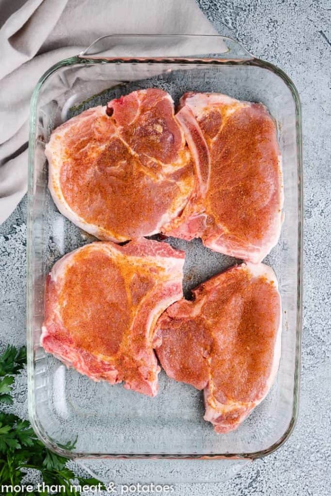 Four pork chops in a pan seasoned with spices.