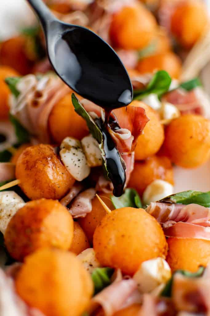 Melon balls, mozzarella, prosciutto, and basil being drizzled with balsamic.