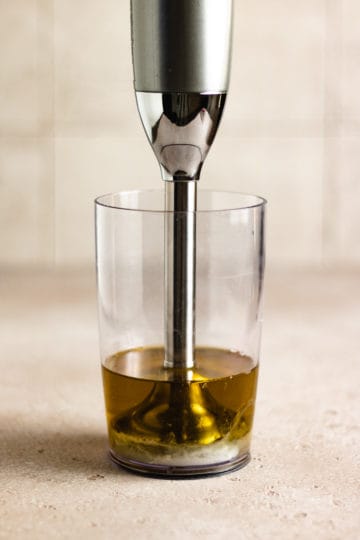 Immersion blender in a plastic cup with oil, egg, and other ingredients.
