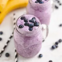 Bananas next to two mugs of blueberry smoothies.