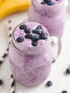 Bananas next to two mugs of blueberry smoothies.