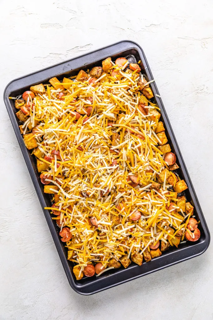 Shredded cheese sprinkled over hot dogs and potatoes on a baking sheet.