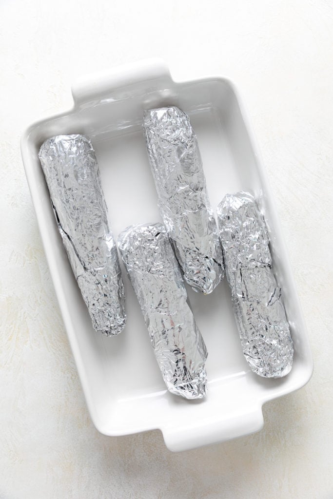 Four ears of corn wrapped in foil and placed in a baking dish.