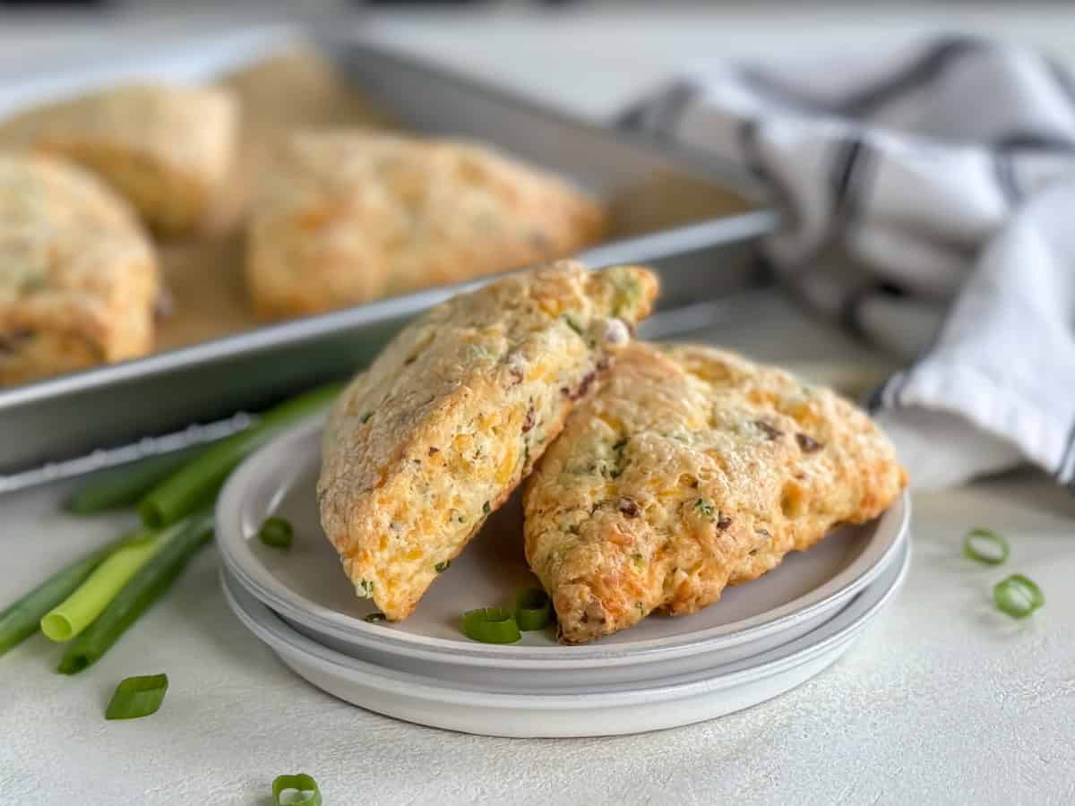 Bacon cheddar scones on a white plate, with green onions as garnish and a tray of scones in the background.