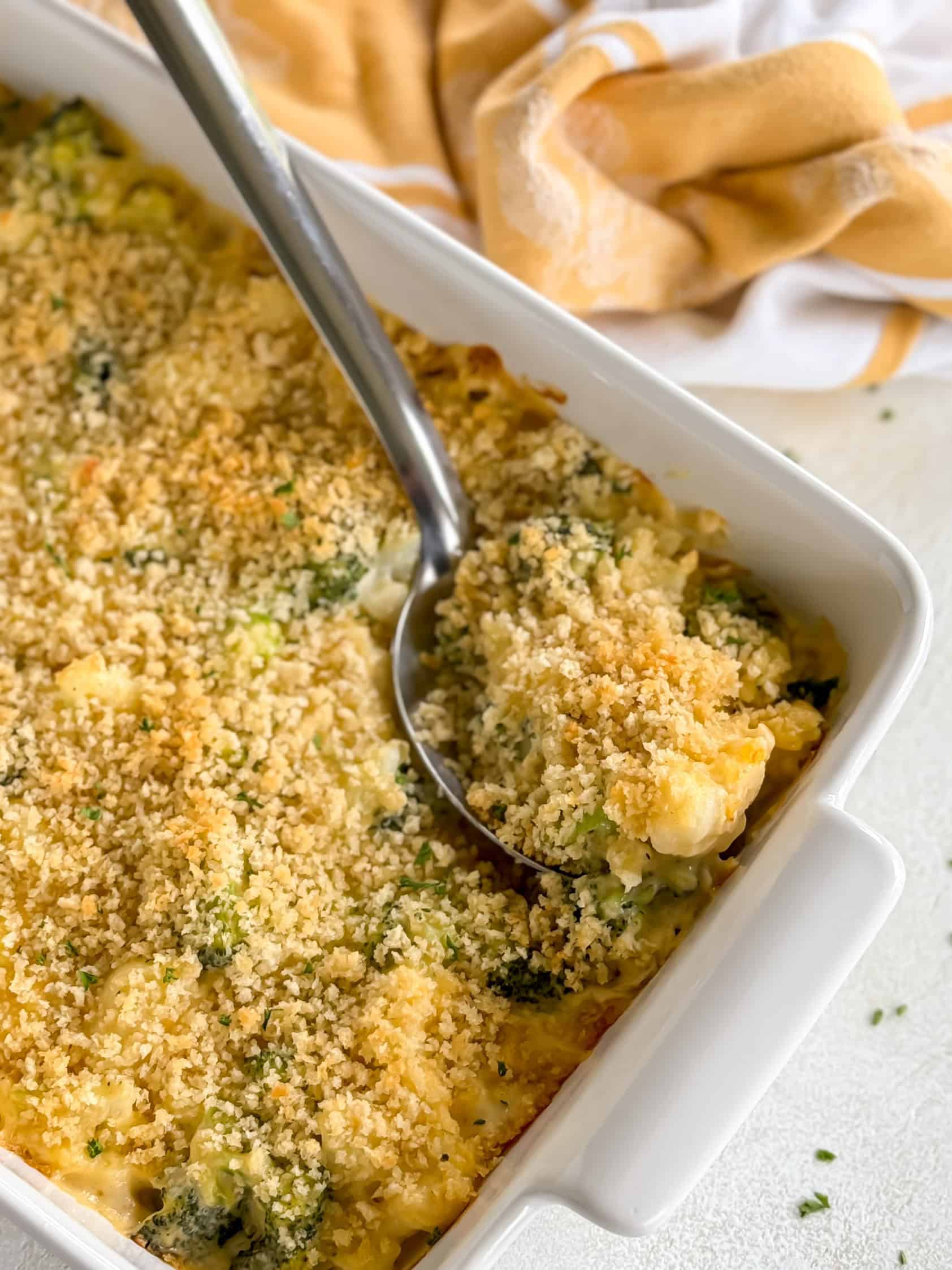 A spoon scooping a serving of cheesy broccoli and cauliflower casserole with a golden breadcrumb topping from a white baking dish.