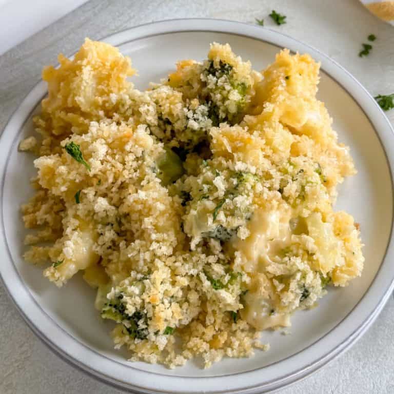 A serving of broccoli and cauliflower casserole with a golden breadcrumb topping on a white plate, with the baking dish in the background.