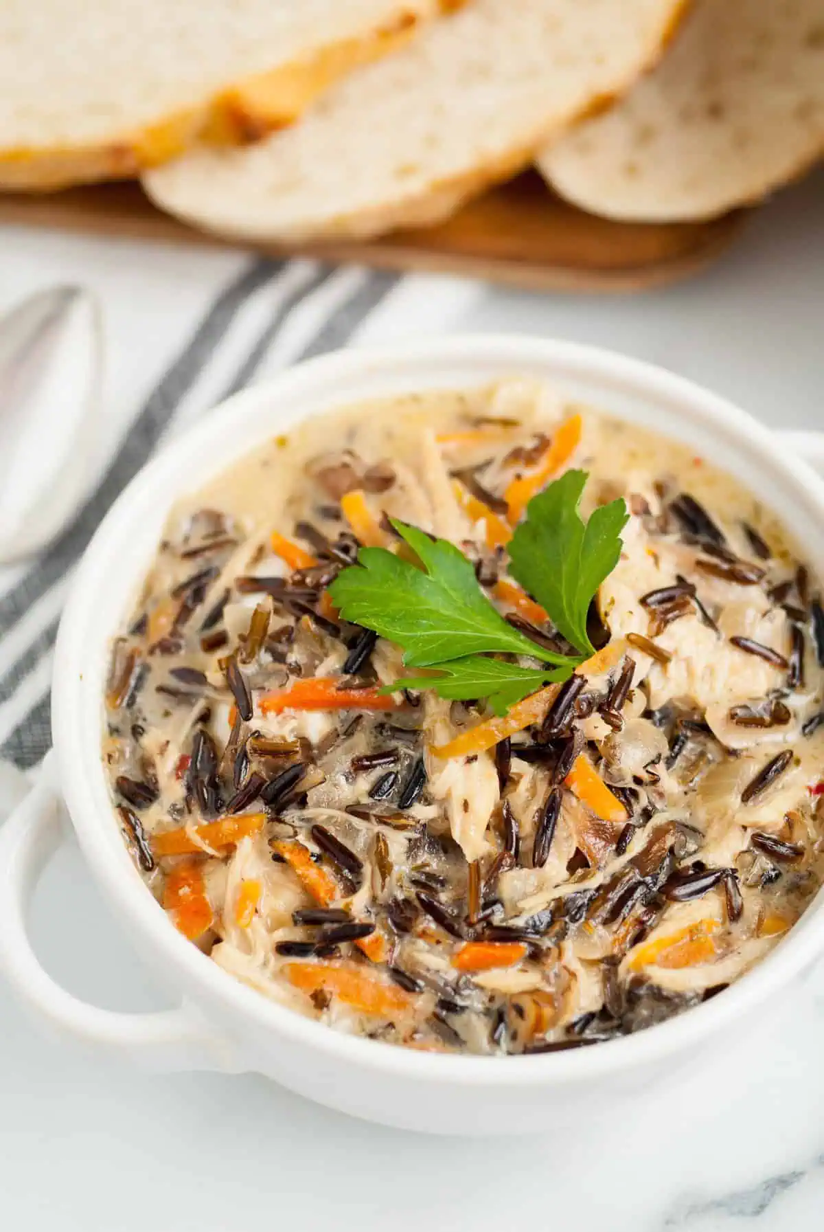 A close-up of a bowl of creamy soup garnished with parsley, showing chunks of chicken and wild rice.