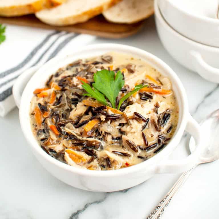 A bowl of creamy chicken wild rice and vegetable soup garnished with a parsley leaf, with sliced bread in the background.