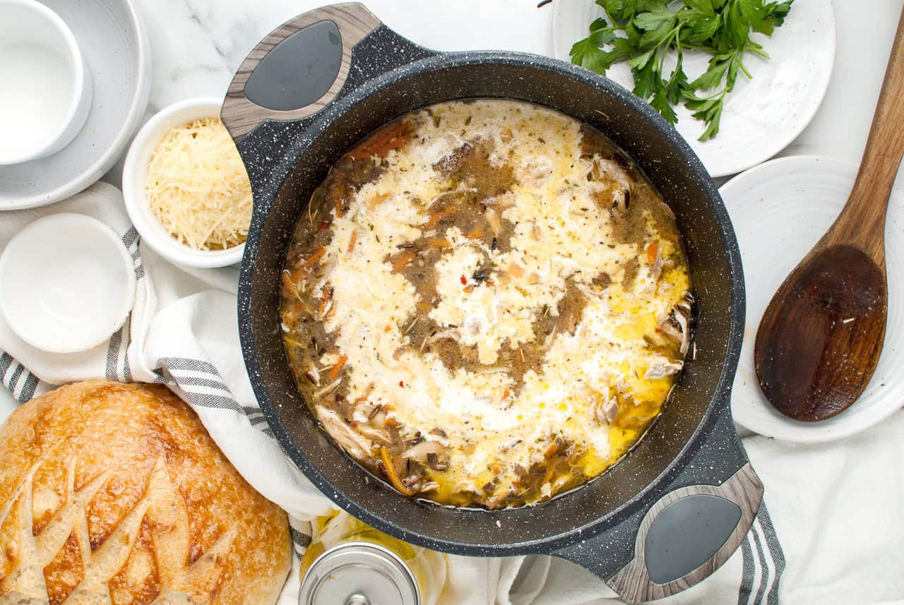 A pot of soup with cream being added, surrounded by ingredients like grated cheese, fresh parsley, and a loaf of bread.