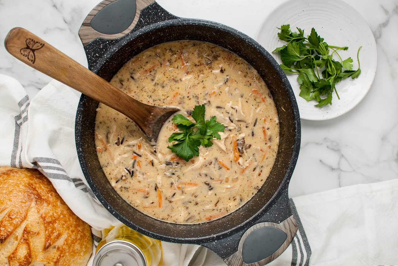 A pot of creamy soup being stirred with a wooden spoon, garnished with parsley, with fresh bread and parsley on the side.