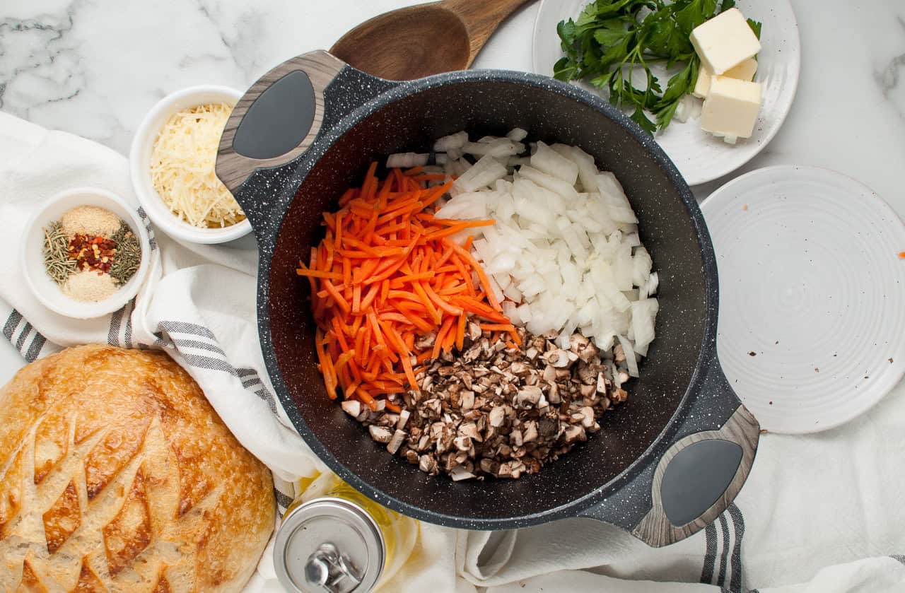 Chopped carrots, onions, and mushrooms in a pot, ready to be cooked, with various ingredients around.