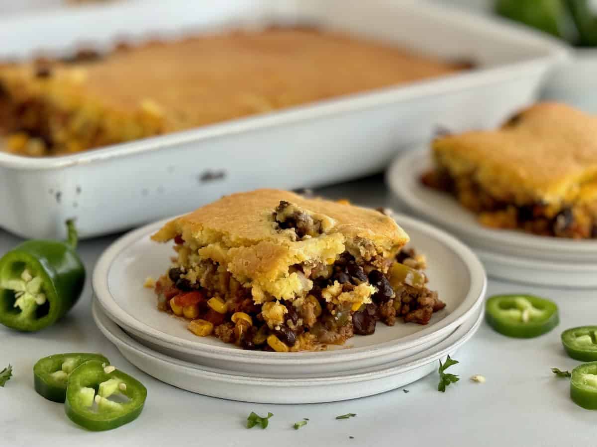 A slice of hamburger cornbread casserole on a white plate, with the baking dish and another slice in the background, garnished with fresh jalapeño slices.