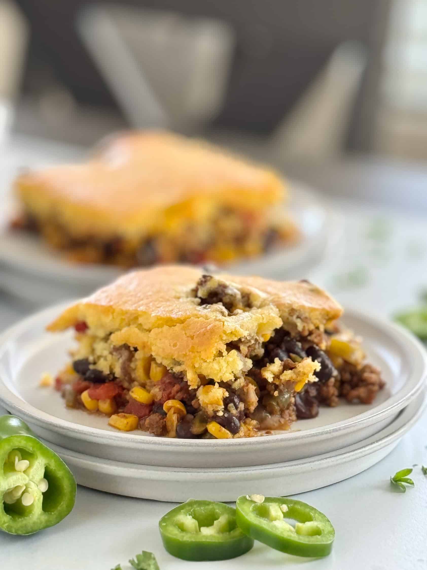 A close-up of a slice of hamburger cornbread casserole on a white plate, showcasing the layers of ground beef, corn, black beans, and cornbread, with the baking dish in the background.