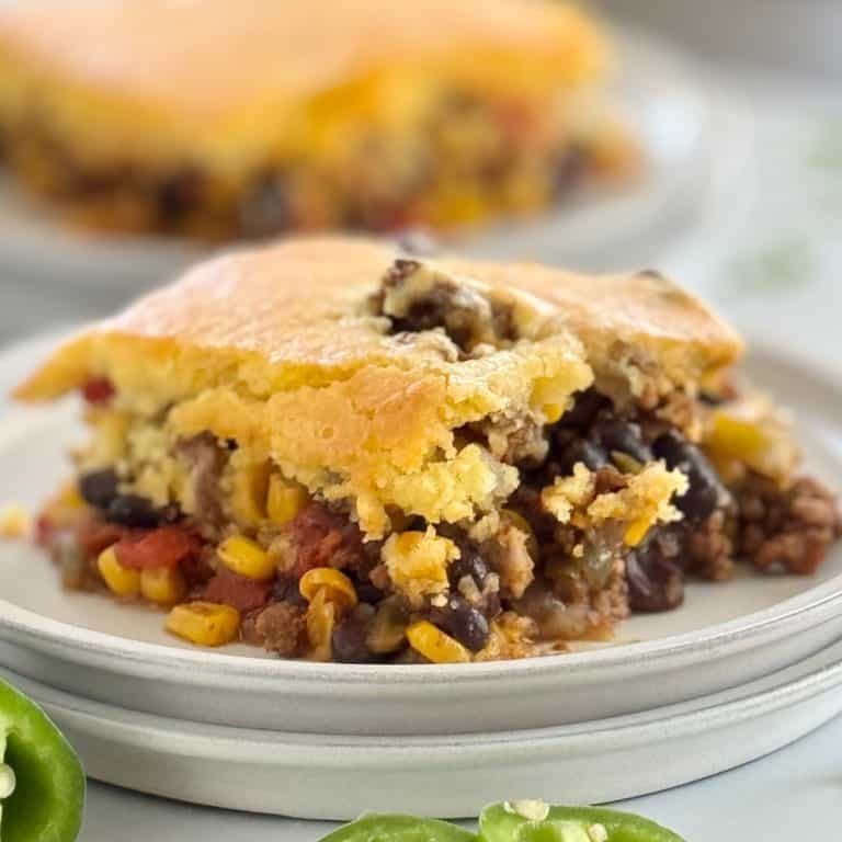 A close-up of a slice of hamburger cornbread casserole on a white plate, showing layers of ground beef, corn, black beans, and cornbread topping.