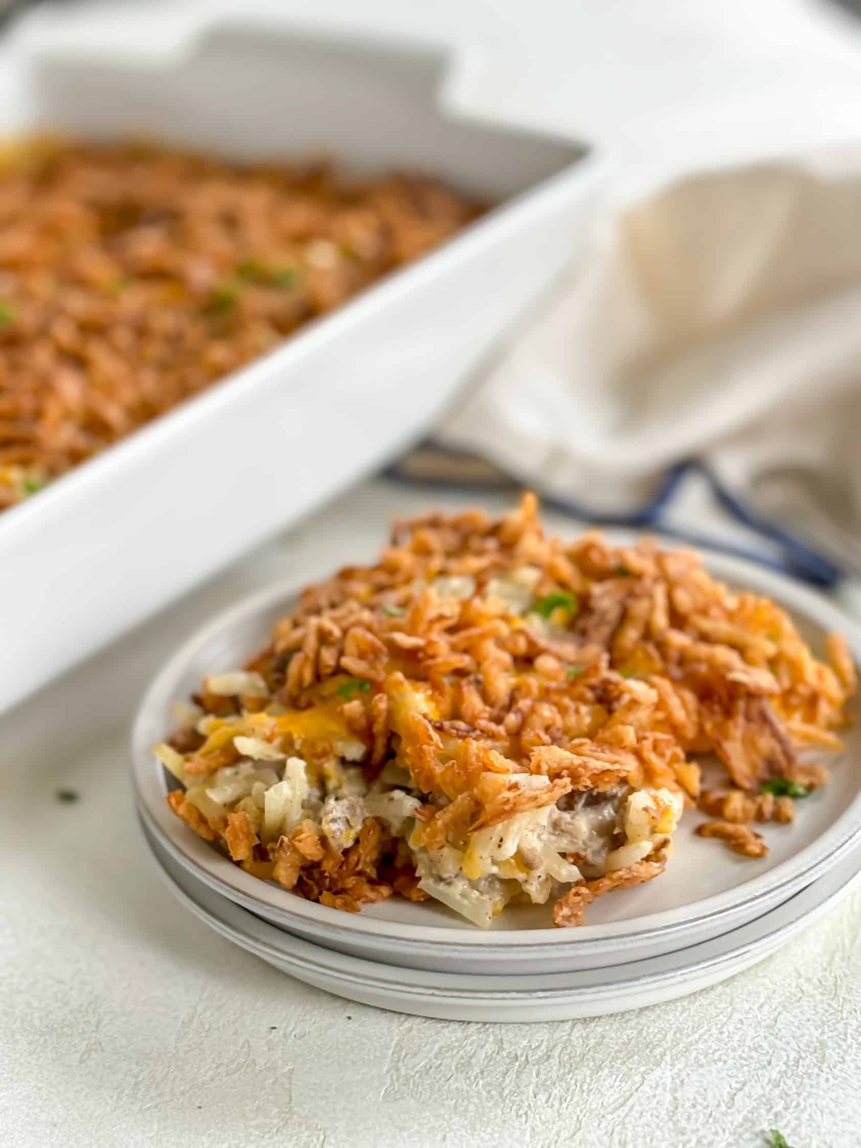 A plate with a serving of hamburger hashbrown casserole in the foreground and the full casserole dish in the background.