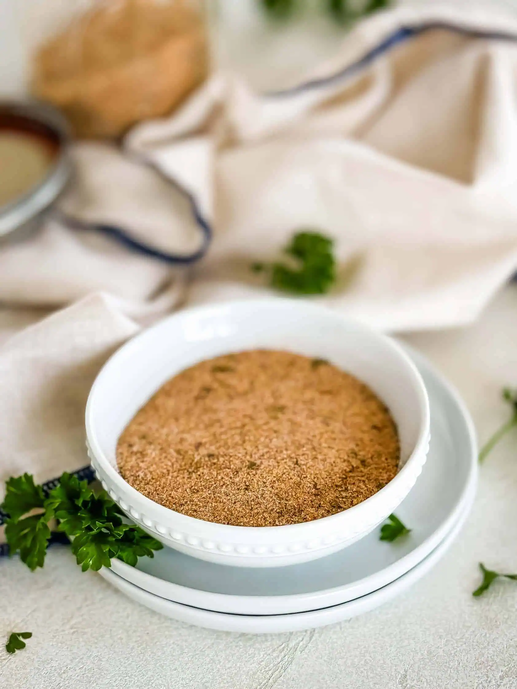 A bowl of homemade seasoned salt on a white plate, surrounded by fresh parsley and a beige cloth napkin.