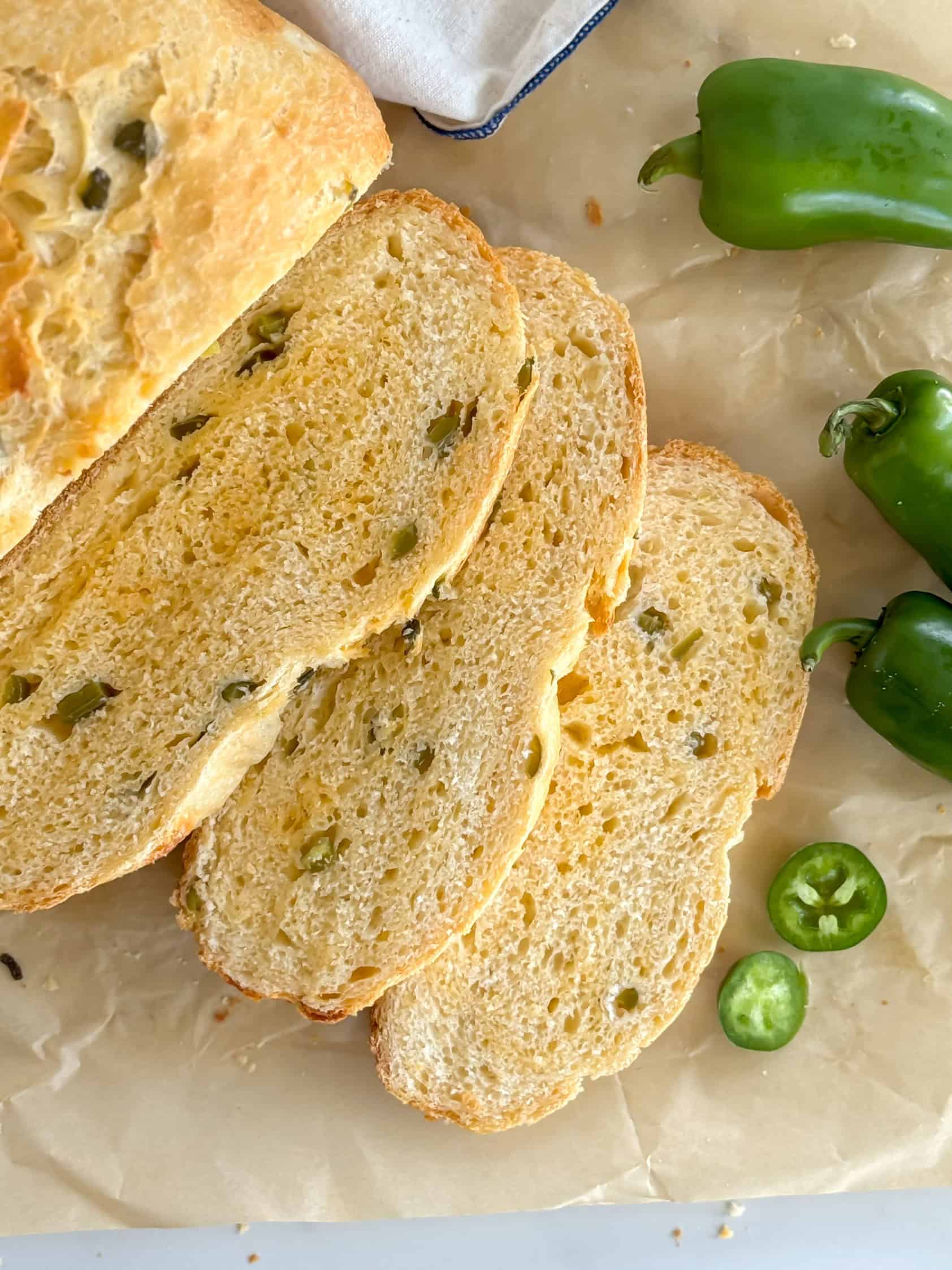 Sliced bread with visible jalapeño and cheese pieces on parchment paper, surrounded by fresh jalapeños.