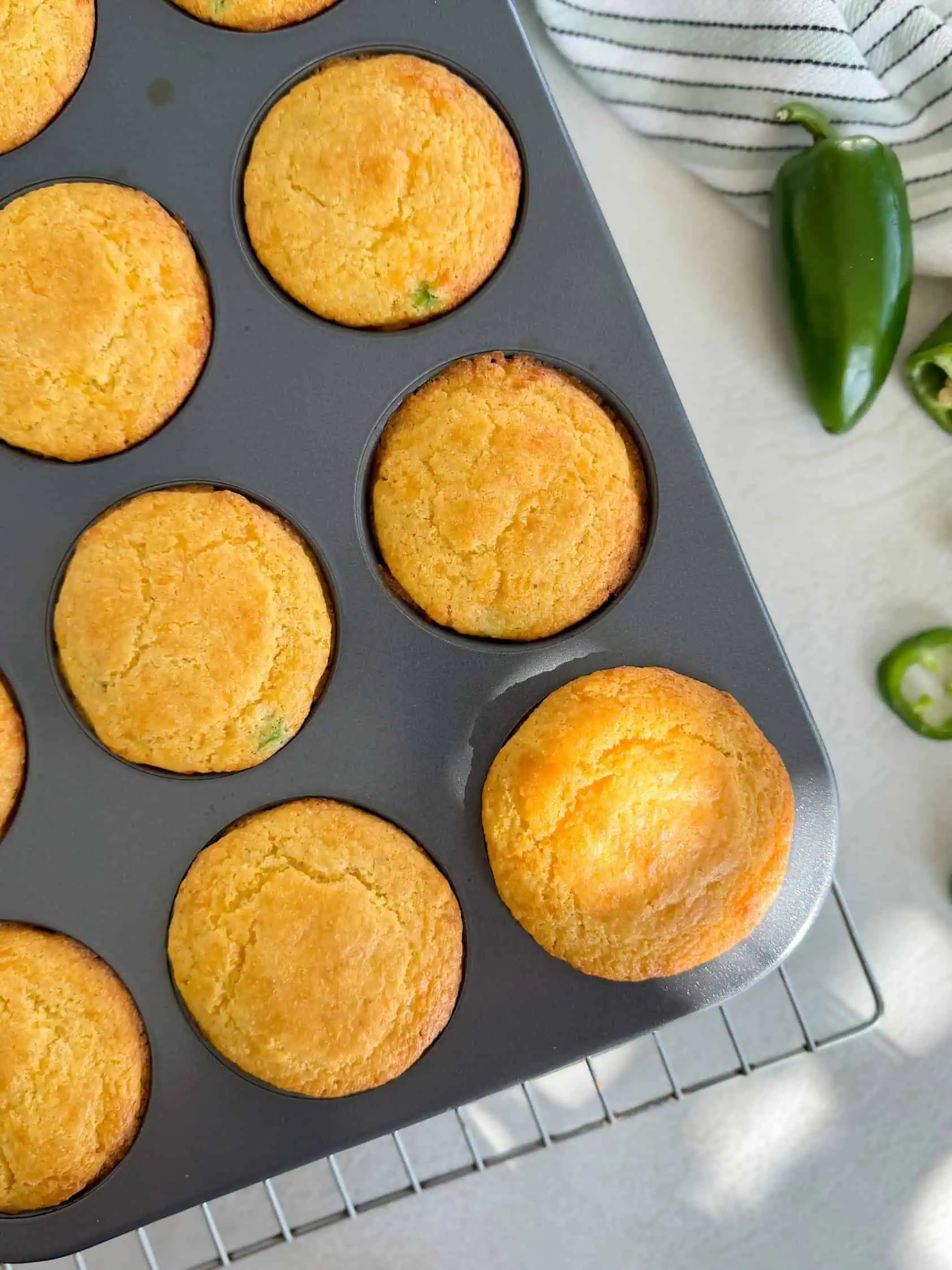 Close-up of a stack of jalapeño cheddar cornbread muffins on a white plate, with fresh jalapeño slices scattered around.