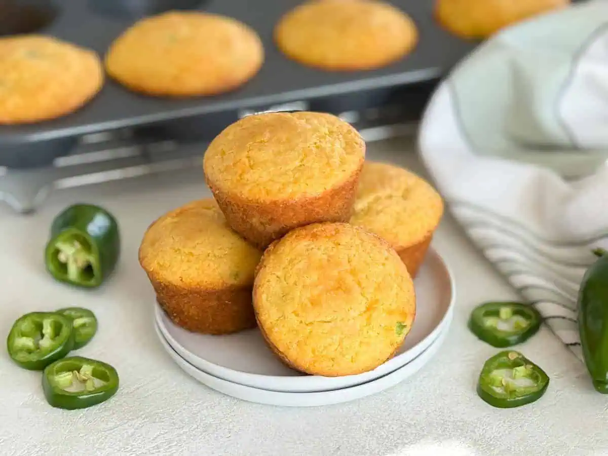 Stack of savory cornbread muffins on a plate, with a muffin tin and fresh jalapeño slices in the background.