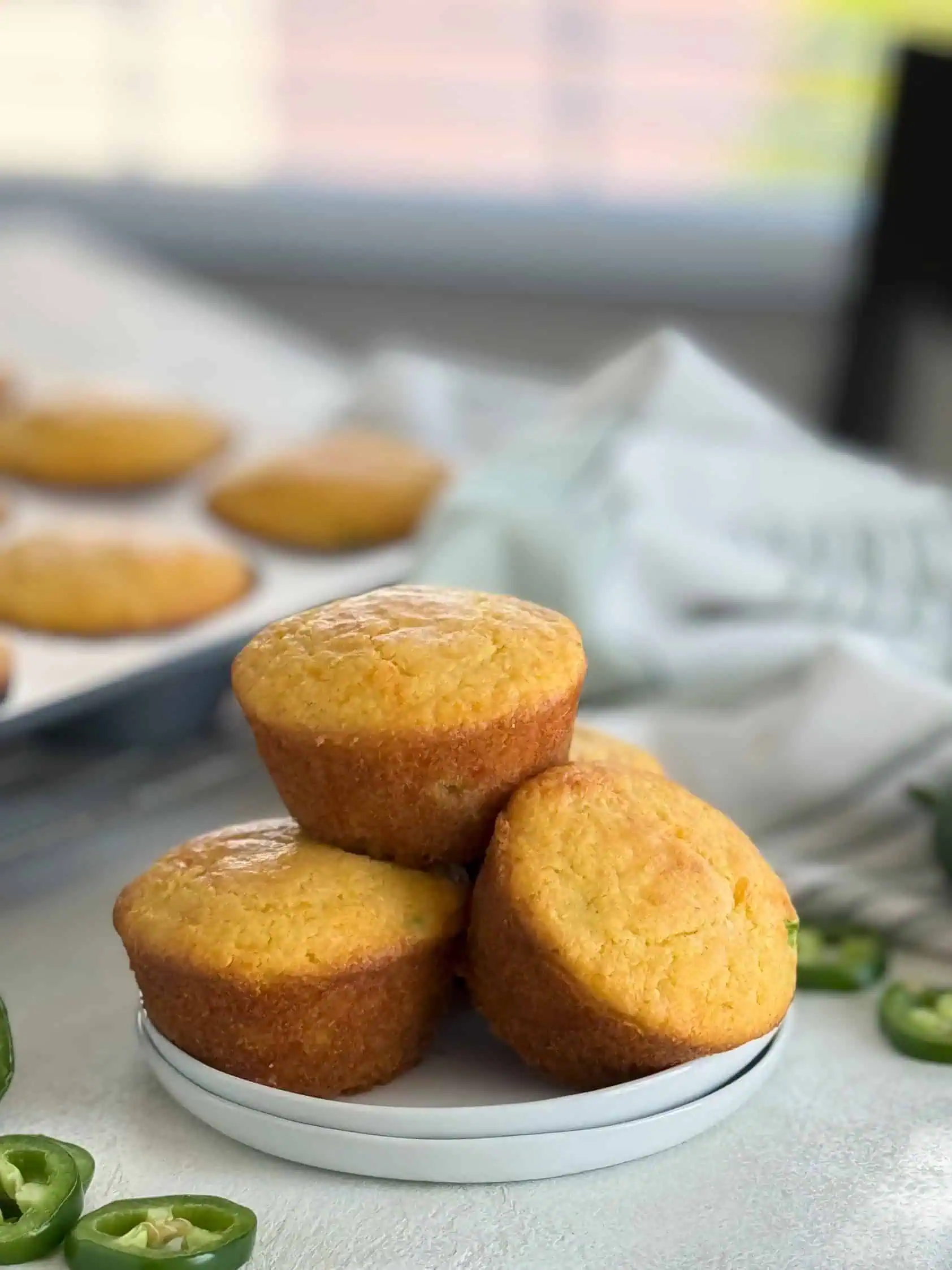 Three cheddar jalapeño muffins stacked on a white plate, with a blurred background of more muffins and jalapeño slices.