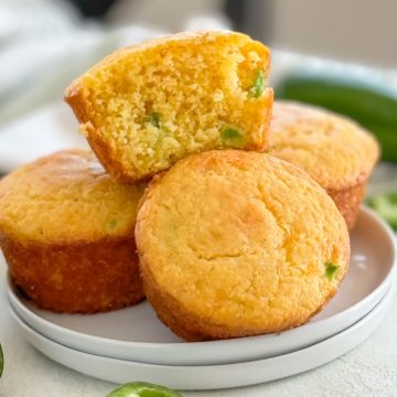 Close-up of a stack of spicy cornbread muffins, one muffin is cut in half showing the moist texture and pieces of jalapeño.