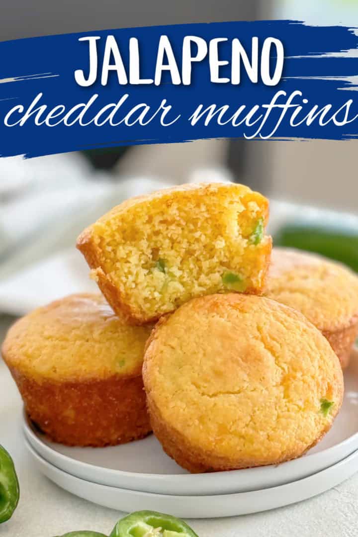 Plate of cheesy jalapeño muffins with one muffin cut in half to show its moist texture, surrounded by fresh jalapeño slices and labeled with 'jalapeño cheddar muffins.