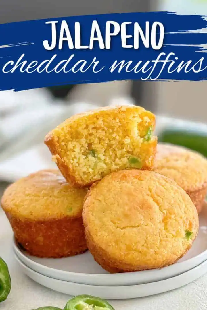 Plate of cheesy jalapeño muffins with one muffin cut in half to show its moist texture, surrounded by fresh jalapeño slices and labeled with 'jalapeño cheddar muffins.