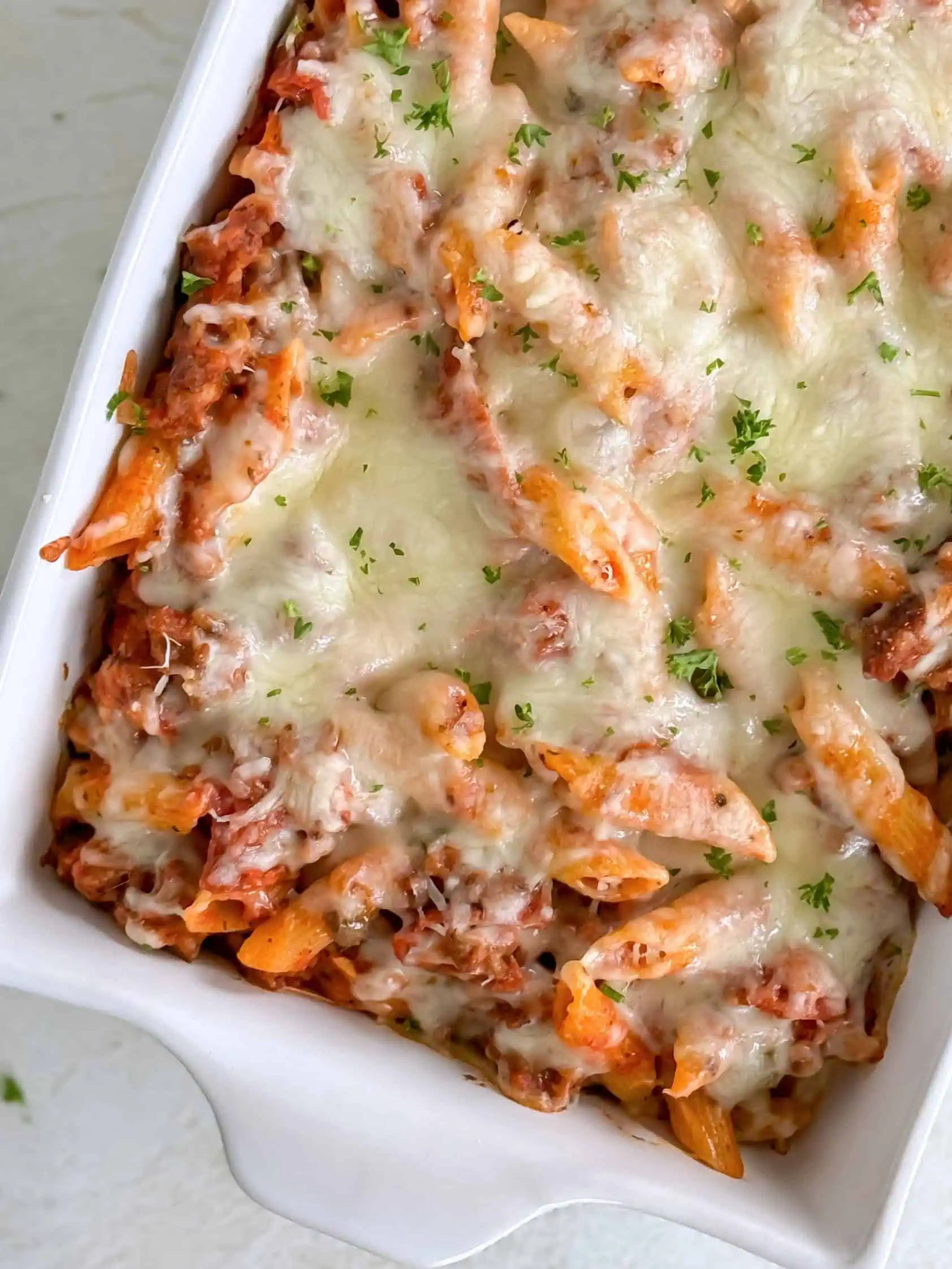 Close-up of a cheesy baked pasta dish with penne, sausage, and melted mozzarella cheese, garnished with chopped parsley in a white baking dish.