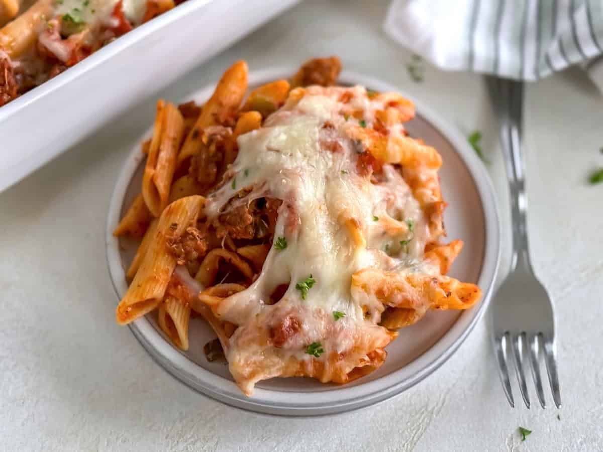 A plate with a generous serving of baked penne pasta topped with melted mozzarella cheese and garnished with parsley, next to a fork on a light background.