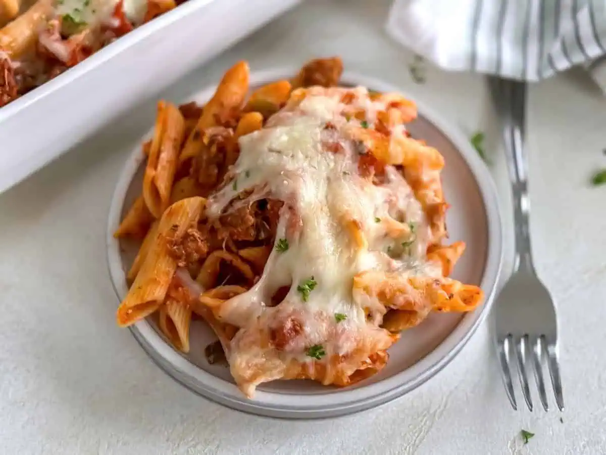 A plate with a generous serving of baked penne pasta topped with melted mozzarella cheese and garnished with parsley, next to a fork on a light background.