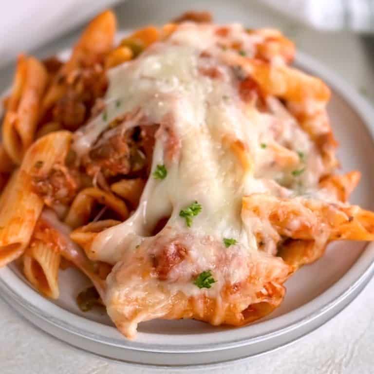 A plate of baked penne pasta with melted cheese, garnished with parsley, with a baking dish of pasta in the background, creating a cozy and inviting presentation.