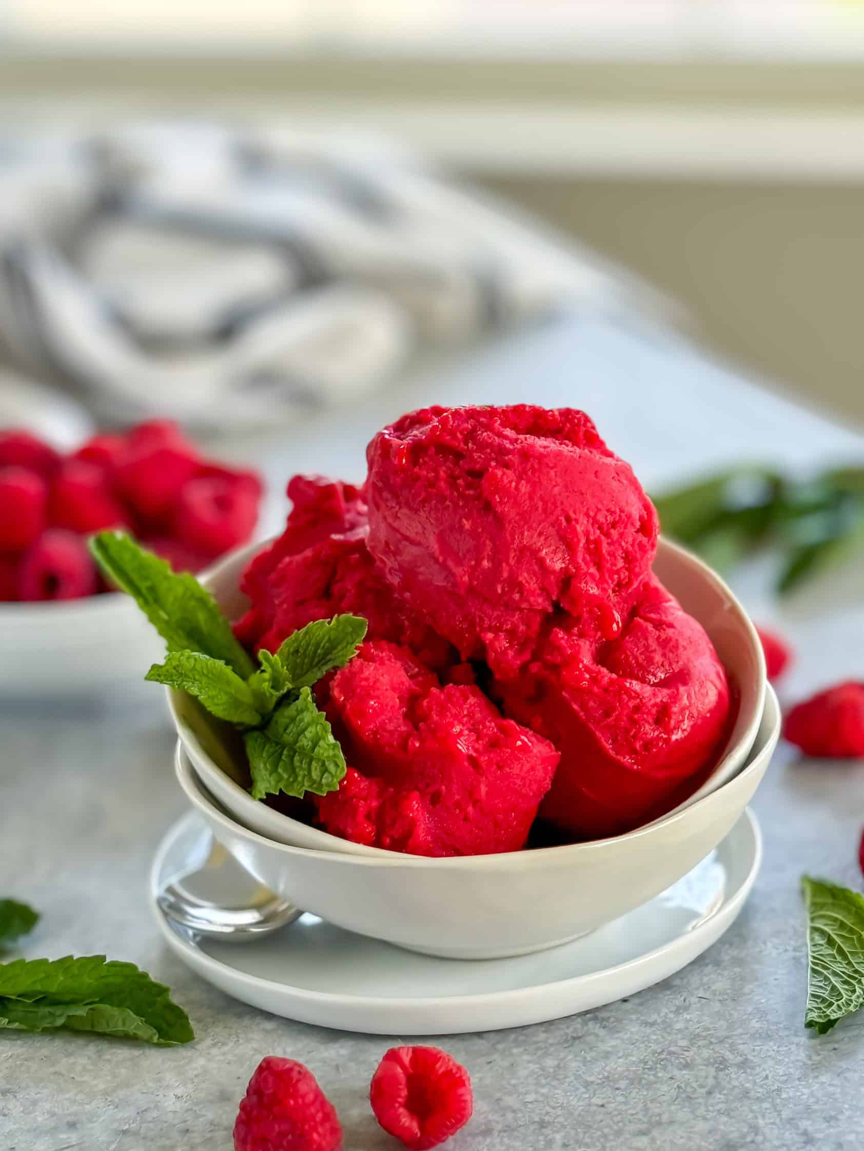 Close-up shot of vibrant raspberry sorbet scoops in a white bowl, garnished with fresh mint leaves, with raspberries in the background.