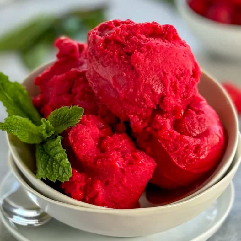 Bowl of raspberry sorbet garnished with fresh mint leaves, with raspberries and mint leaves on a light gray surface.