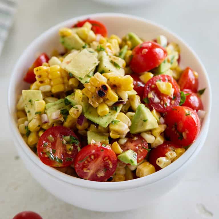 A close-up of the finished salad in a white bowl, showcasing grilled corn, halved cherry tomatoes, diced avocado, red onion, and cilantro.
