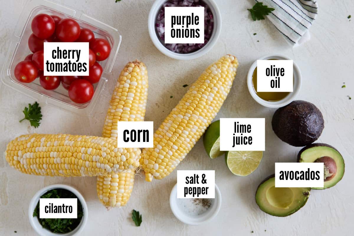 Fresh ingredients laid out on a counter including cherry tomatoes, corn on the cob, purple onions, avocados, lime juice, olive oil, cilantro, and salt & pepper.