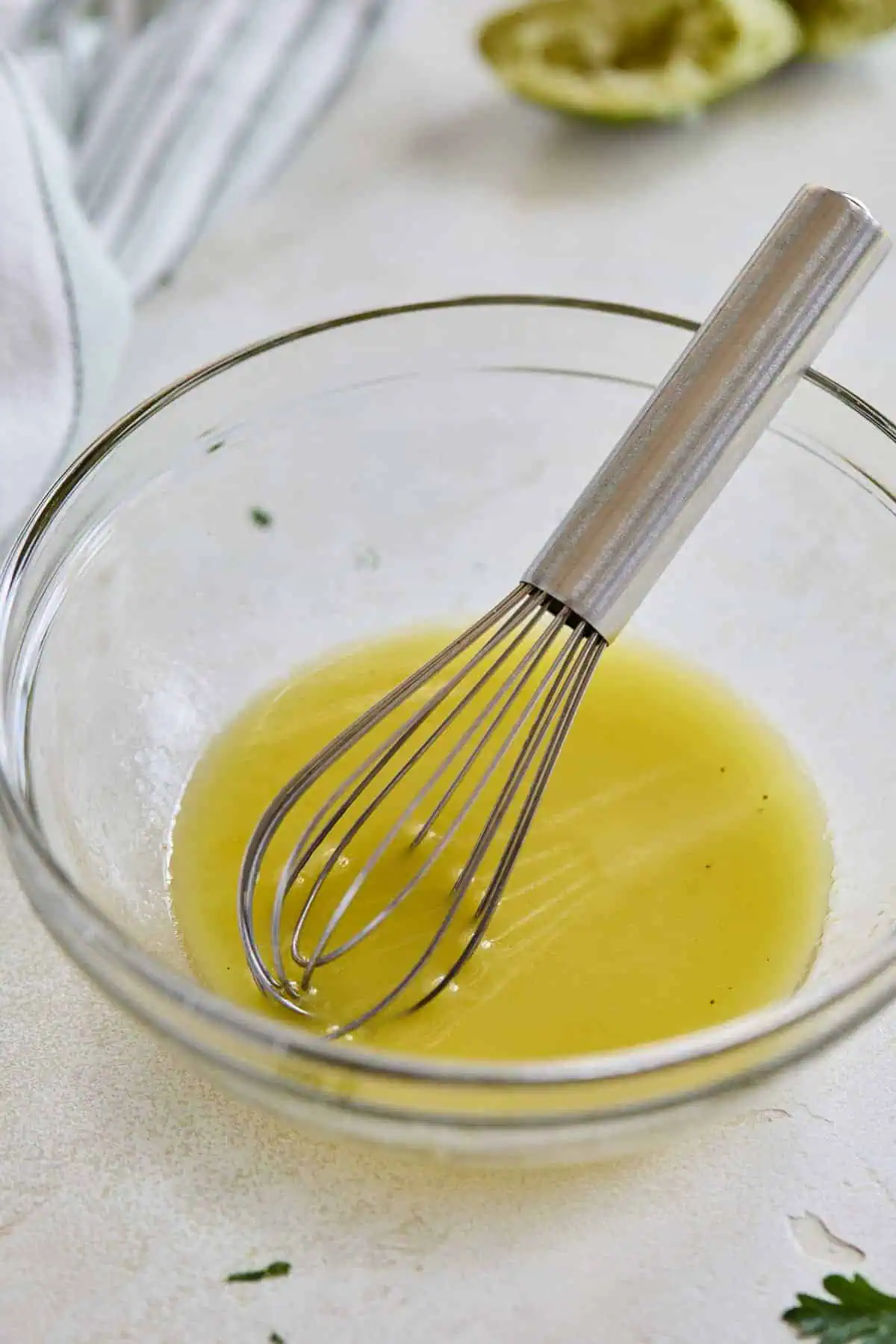 A glass bowl with a freshly whisked dressing, featuring a metal whisk and squeezed lime halves in the background.