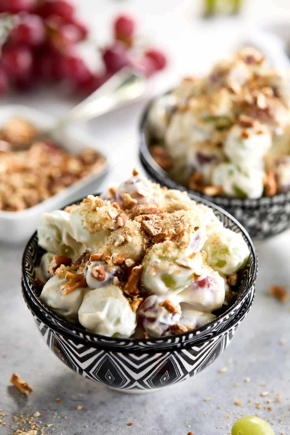 A bowl of grape salad topped with chopped pecans and crushed pretzels, with grapes in the background.