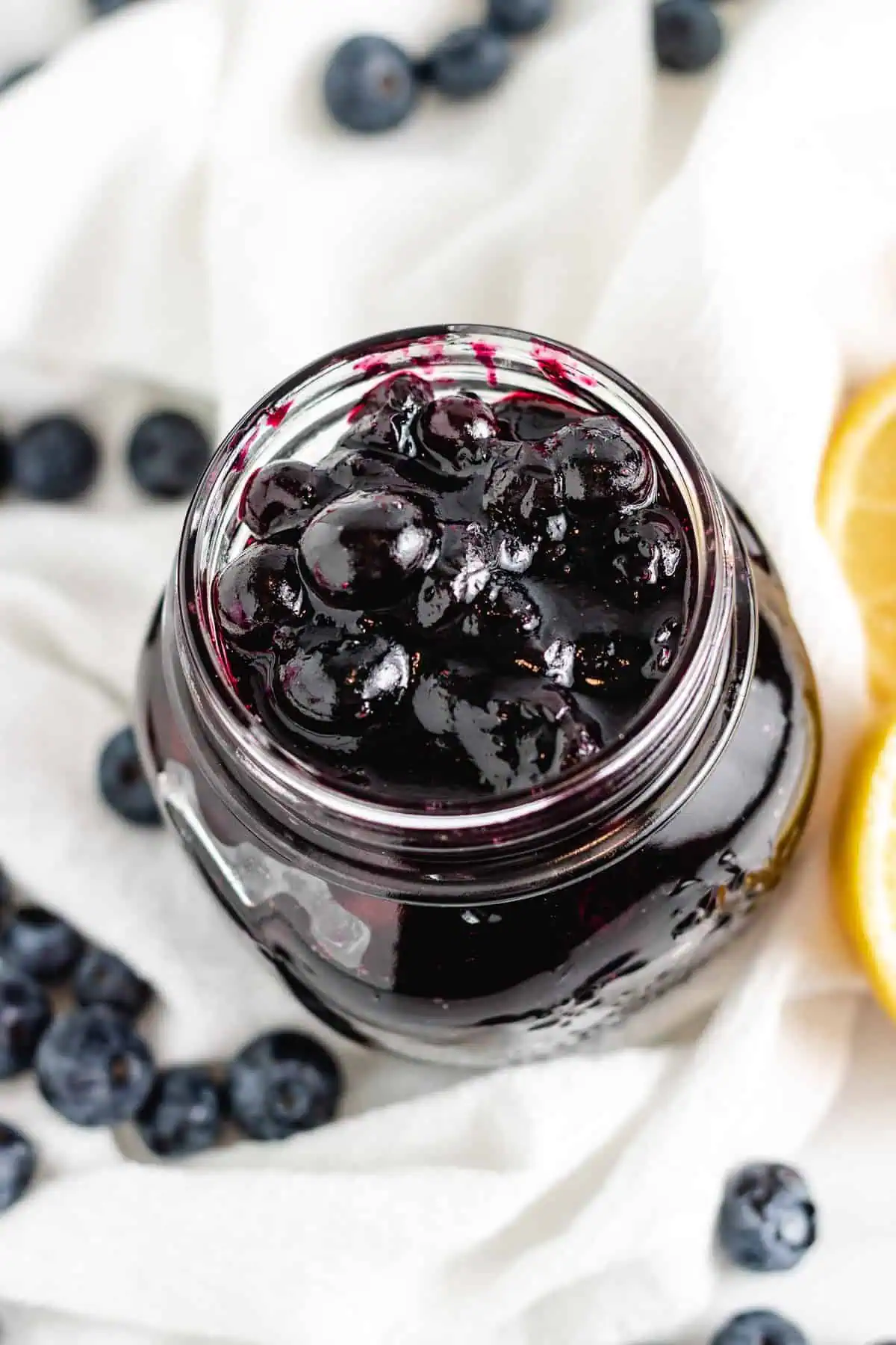 A top-down view of a jar filled with rich blueberry compote, with fresh blueberries scattered around and lemon slices on the side.