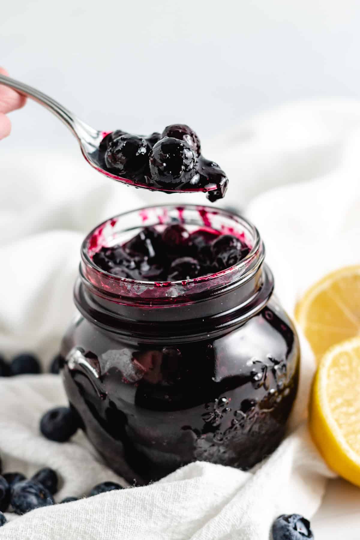 A spoonful of thick blueberry compote being lifted from a jar, surrounded by fresh blueberries and lemon slices.