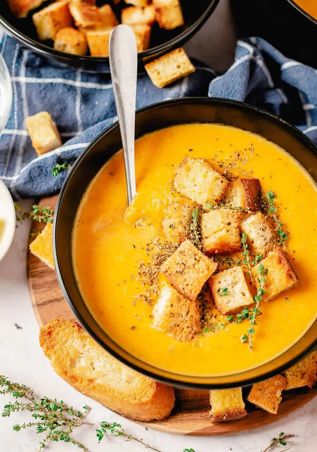 Butternut squash and sweet potato soup in a black bowl, topped with croutons, fresh thyme, and a spoon, with bread slices and a blue cloth in the background.