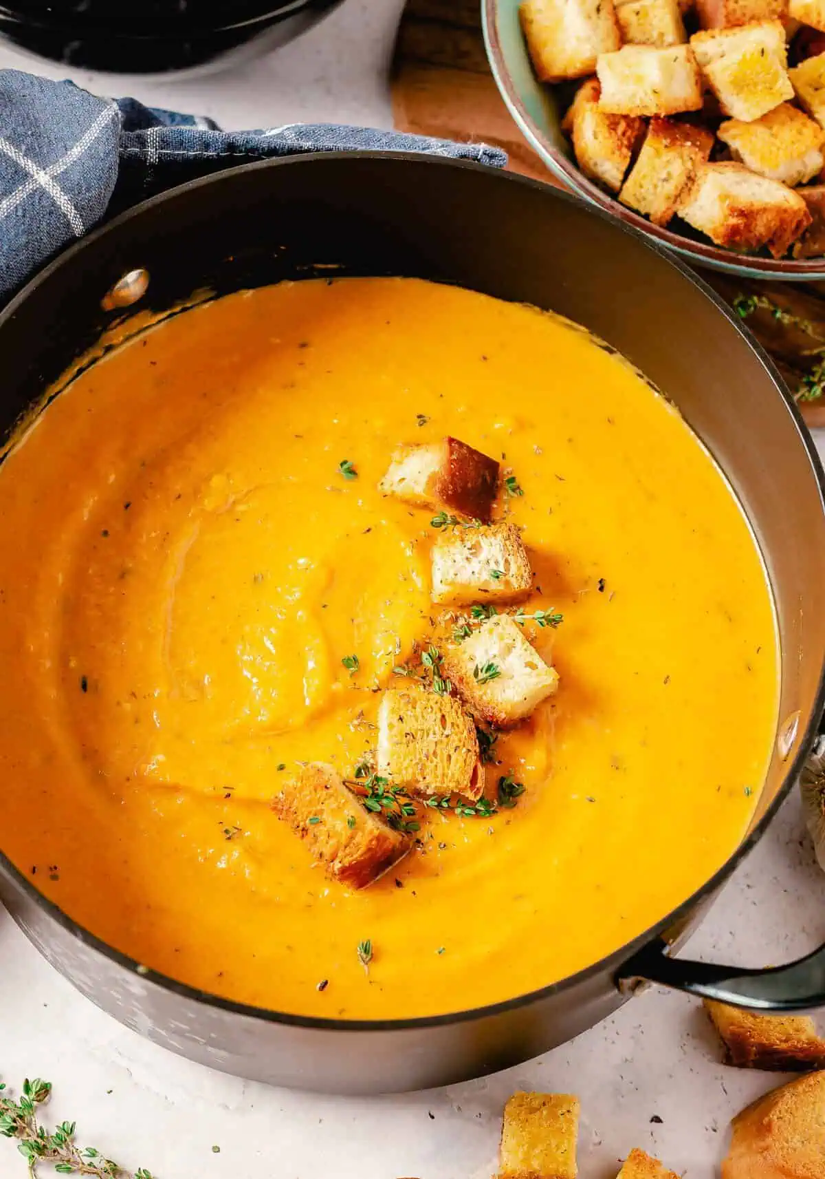 A pot of smooth butternut squash and sweet potato soup with croutons and fresh thyme on top, next to a small bowl of additional croutons.