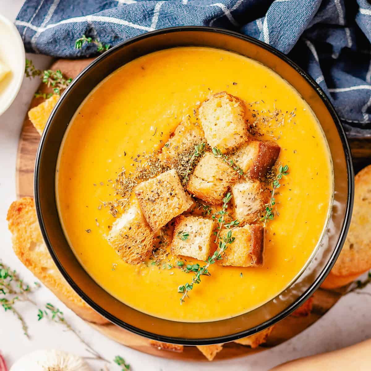 Overhead view of a bowl of butternut squash and sweet potato soup, garnished with croutons and thyme, placed on a wooden cutting board with slices of bread.