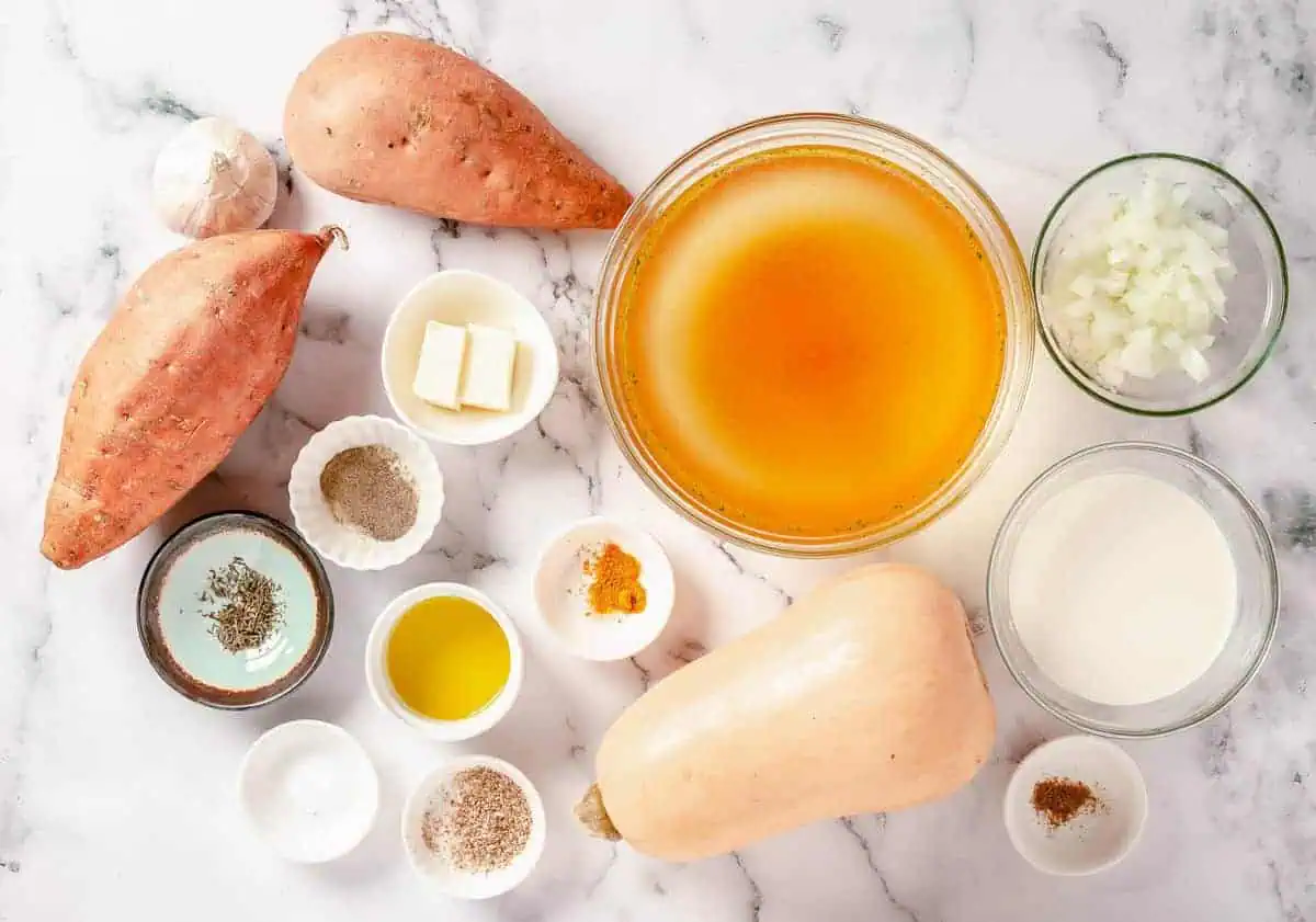 Ingredients for butternut squash and sweet potato soup laid out on a marble countertop, including sweet potatoes, butternut squash, broth, and seasonings.