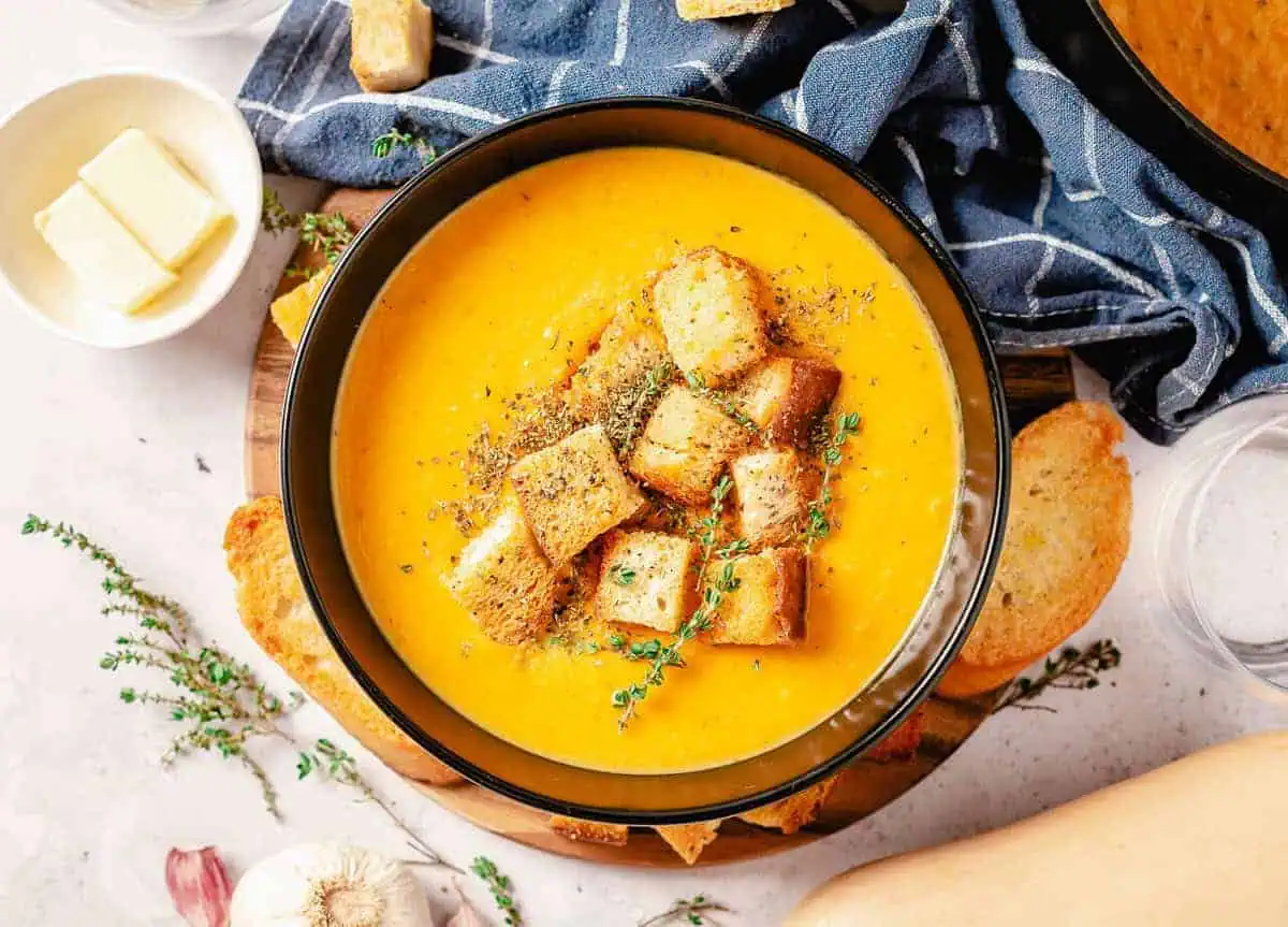 A bowl of creamy butternut squash soup topped with golden croutons and fresh thyme, with a blue cloth napkin and garlic bulb in the background.