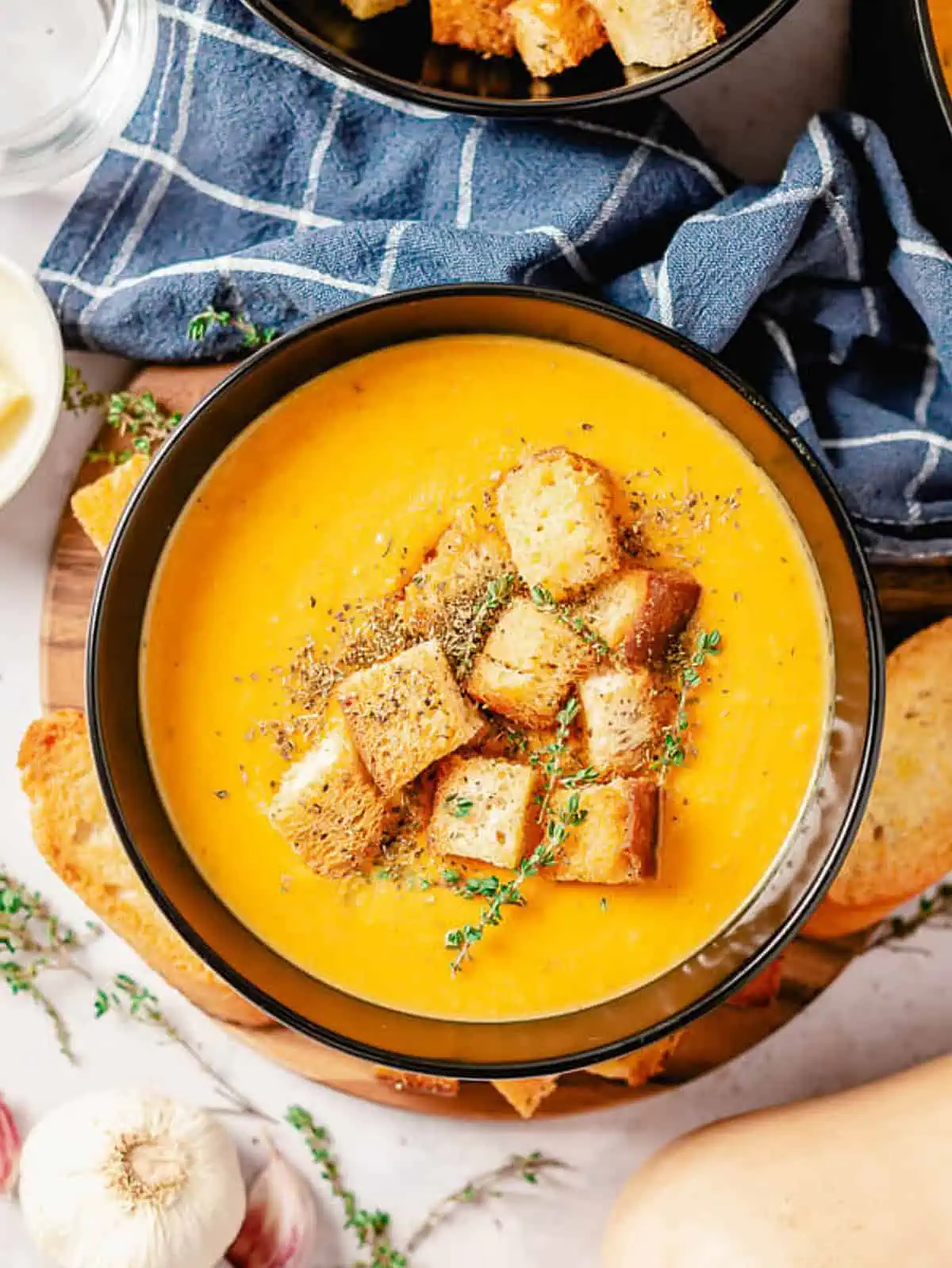 Butternut squash and sweet potato soup in a black bowl, topped with crispy croutons and a sprig of thyme, next to a bowl of additional croutons.