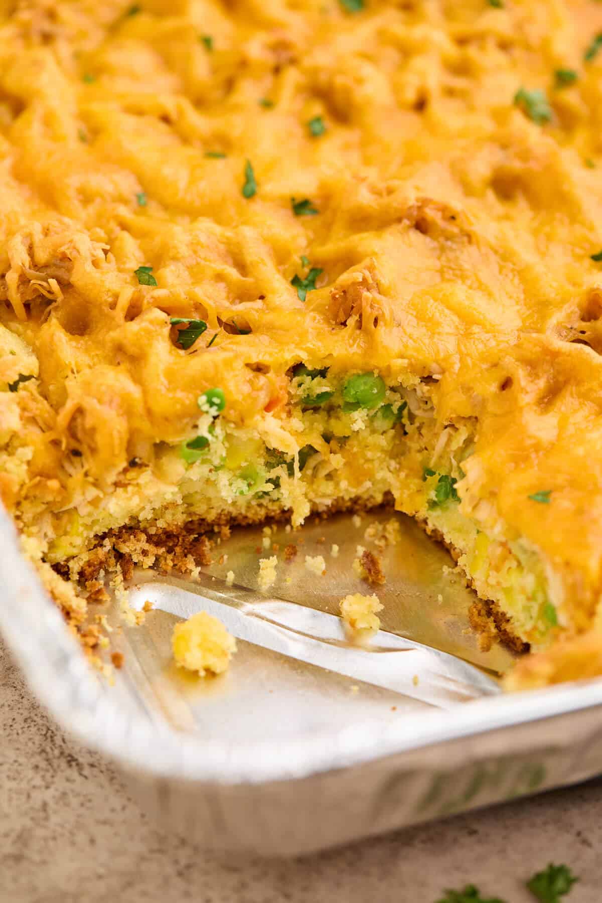 A close-up of the chicken cornbread casserole in a baking dish with a serving removed, revealing the layers of cornbread, vegetables, chicken, and melted cheddar cheese.