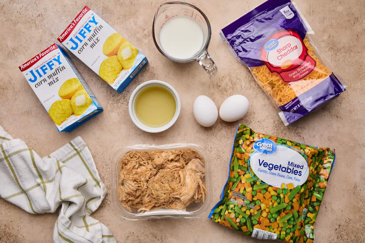 All the ingredients for chicken cornbread casserole displayed on a countertop, including jiffy corn muffin mix, milk, eggs, chicken broth, mixed vegetables, and shredded cheddar cheese.