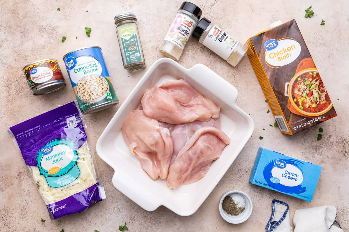 Ingredients for crock pot white chicken chili laid out on a beige surface, including chicken breasts, cannellini beans, green chiles, chicken broth, cream cheese, shredded monterey jack cheese, and seasonings.