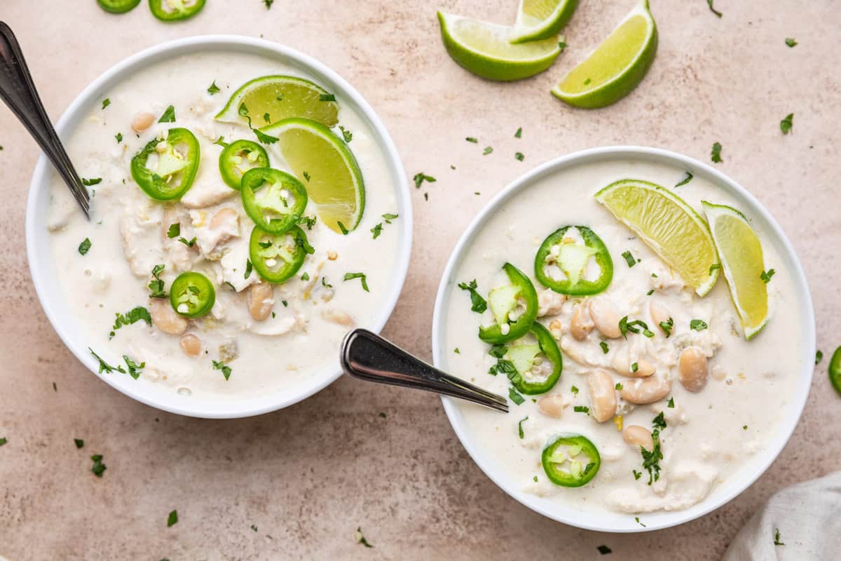 Two bowls of creamy slow cooker white chicken chili garnished with fresh jalapeños and lime slices.