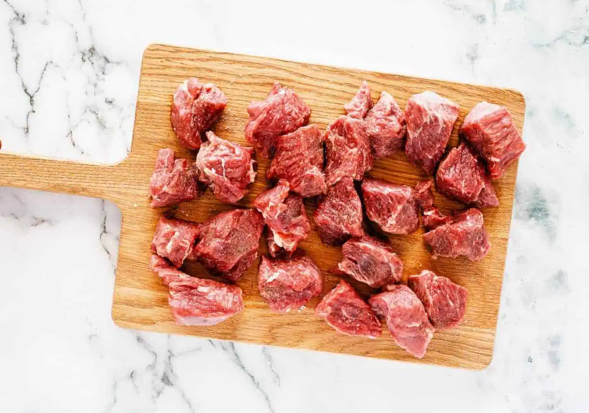 Raw beef chunks seasoned with salt and pepper on a wooden cutting board.